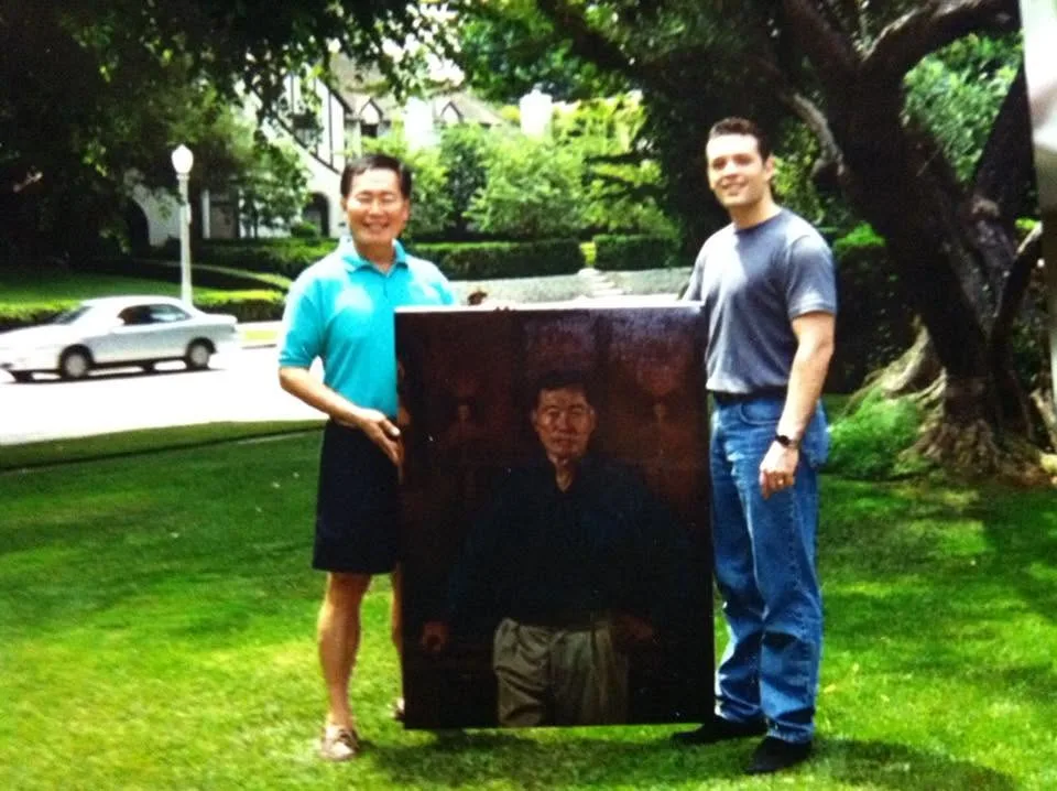 A young Kevin Murphy and George Takei holding a commissioned portrait on a lawn